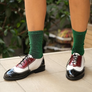 May include: Black and white leather oxford shoes with a burgundy accent. The shoes have laces and are worn with green sparkly socks.