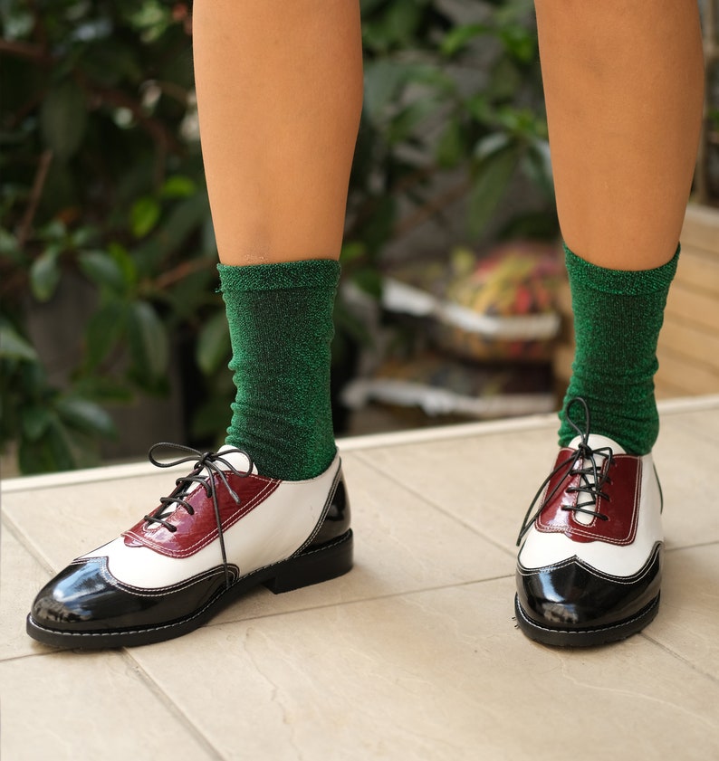 May include: Black and white leather oxford shoes with a burgundy accent. The shoes have laces and are worn with green sparkly socks.