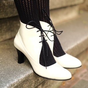 May include: White leather ankle boots with black lace-up detailing. The boots have a high heel and are worn with black fishnet stockings.