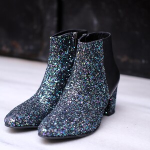 May include: A pair of black ankle boots covered in iridescent glitter. The boots have a pointed toe, a side zipper, and a block heel. The back of the boot is black leather. The boots are set against a dark background.