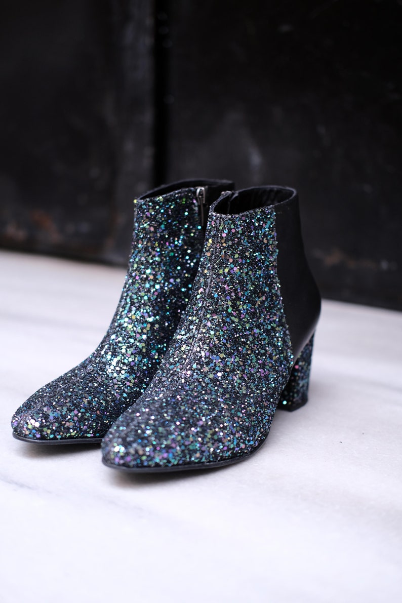 May include: A pair of black ankle boots covered in iridescent glitter. The boots have a pointed toe, a side zipper, and a block heel. The back of the boot is black leather. The boots are set against a dark background.