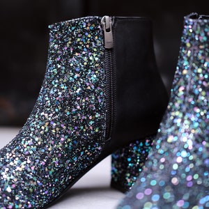 May include: Close-up of two glitter-covered ankle boots. The boots have a black base with iridescent glitter, a side zipper, and a block heel. The zipper pull is silver and has the letters "YKK" on it. The boots are on a white surface.