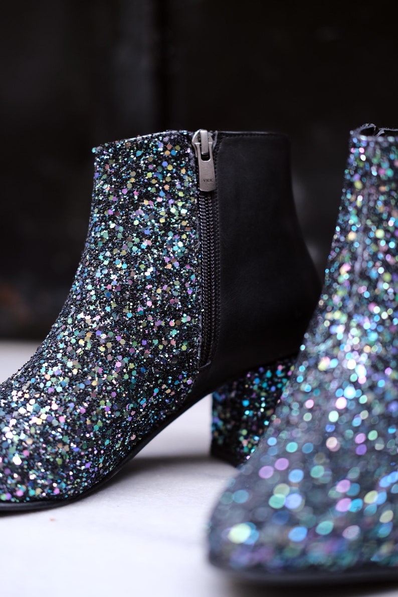 May include: Close-up of two glitter-covered ankle boots. The boots have a black base with iridescent glitter, a side zipper, and a block heel. The zipper pull is silver and has the letters "YKK" on it. The boots are on a white surface.