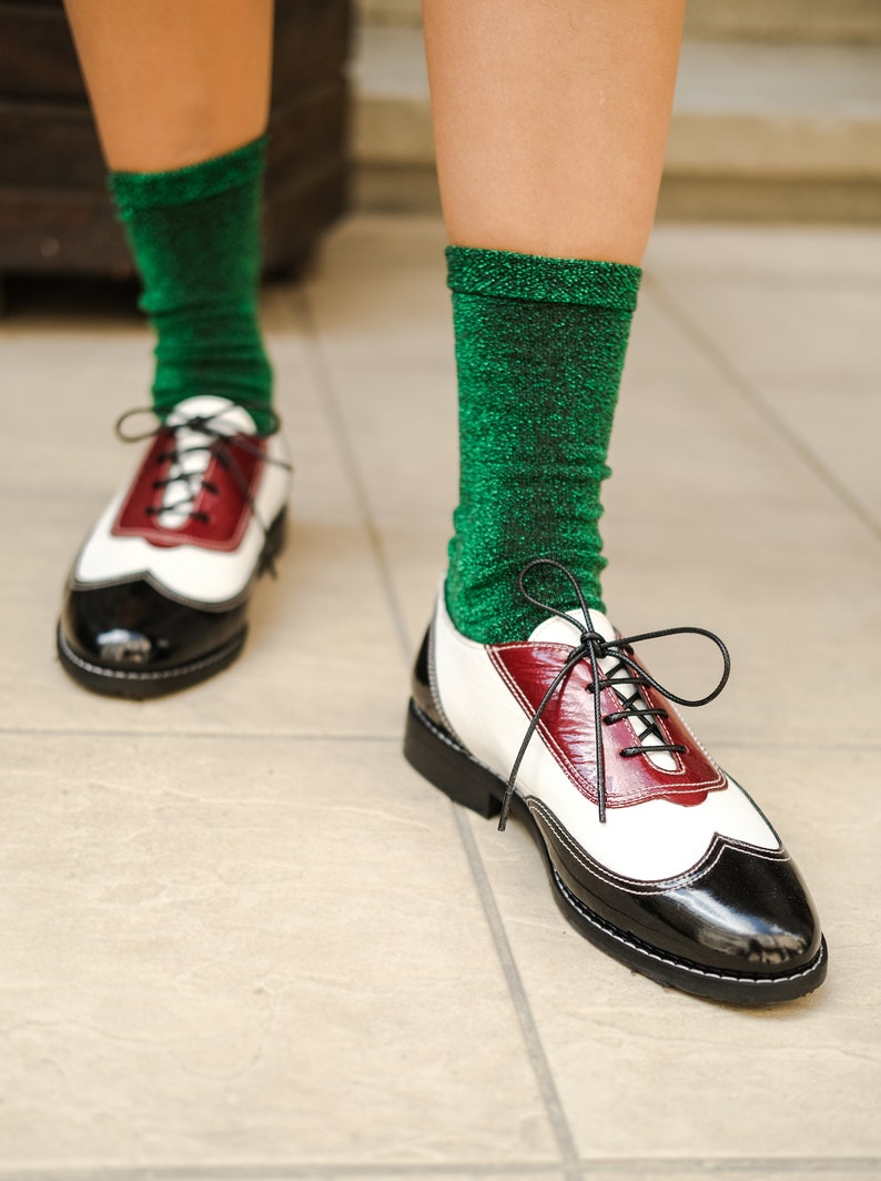 May include: A pair of white and black leather shoes with red accents. The shoes have laces and are worn with green sparkly socks.