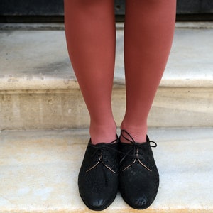 May include: A pair of black leather lace-up shoes with a gold trim on the laces. The shoes are worn by a person with red tights.