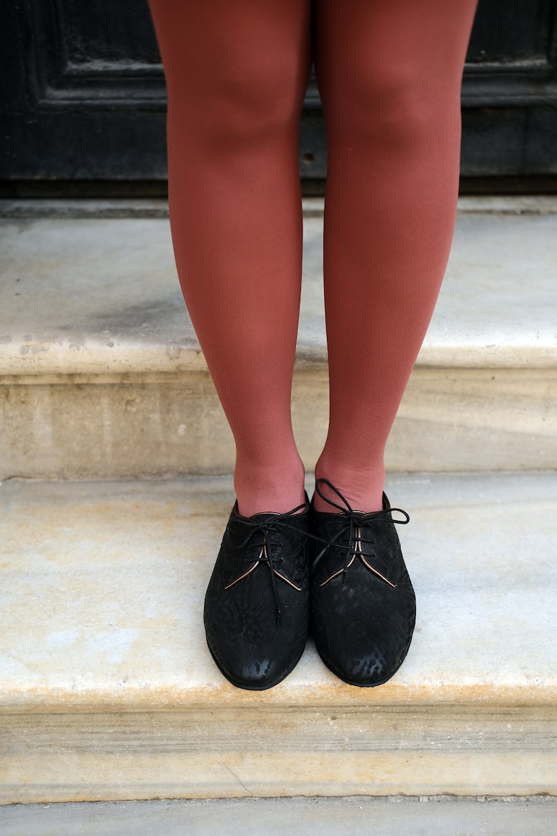 May include: A pair of black leather lace-up shoes with a gold trim on the laces. The shoes are worn by a person with red tights.