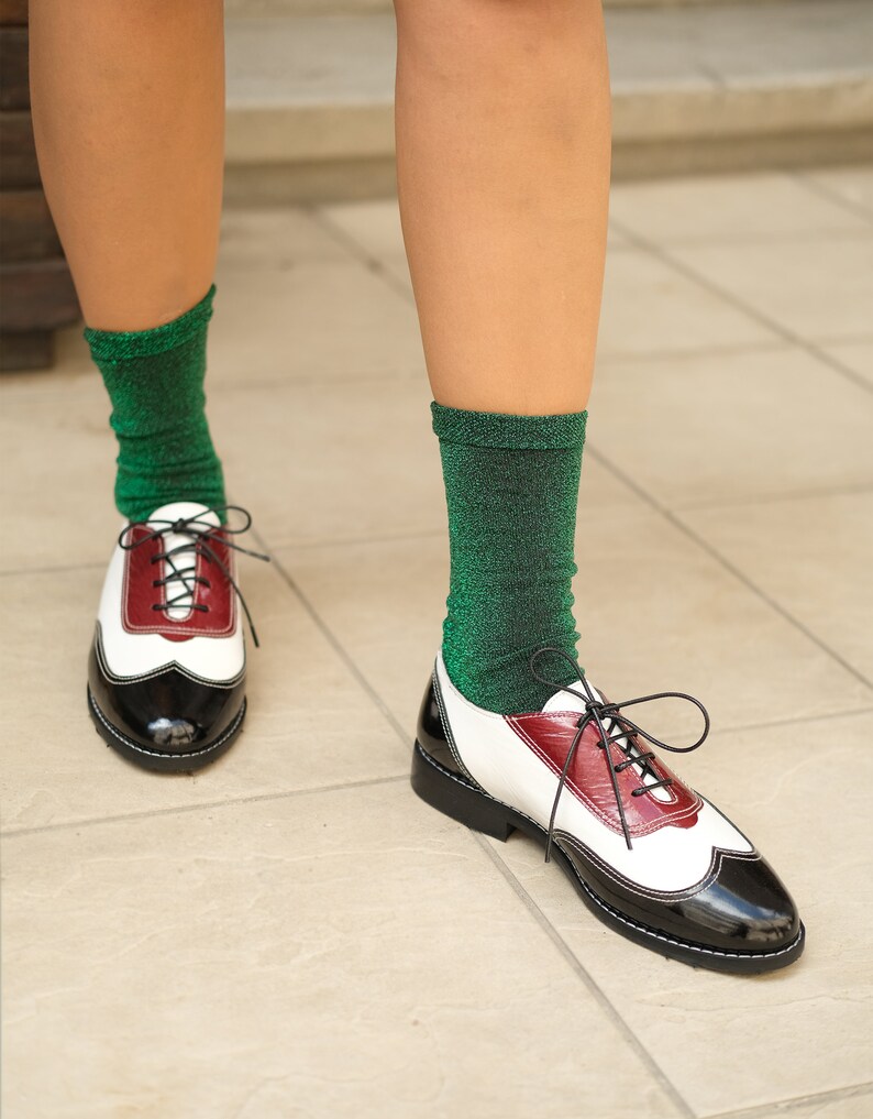 May include: A pair of black and white leather oxfords with red accents. The shoes have laces and a pointed toe. The person wearing the shoes is wearing green sparkly socks.