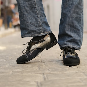 Könnte beinhalten: Ein Paar schwarze Lederschuhe mit silbernem Streifen-Detail. Die Schuhe haben einen Schnürverschluss und eine abgerundete Spitze.