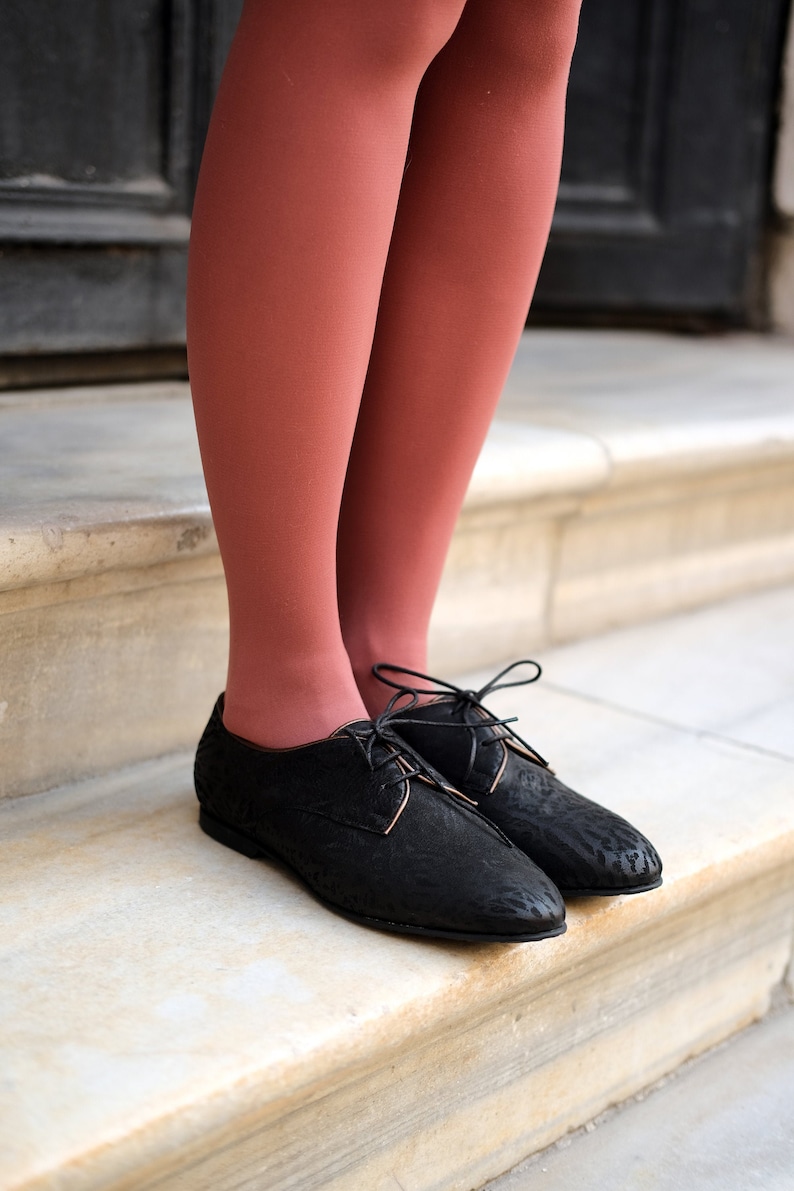 May include: A pair of black lace-up shoes with a low heel. The shoes are being worn by someone wearing rust-coloured tights.