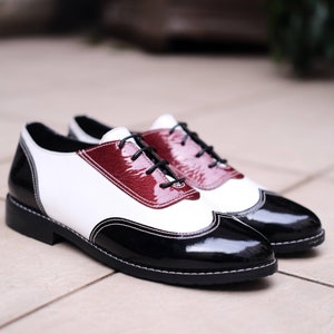 May include: A pair of black and white leather shoes with a burgundy accent. The shoes have a lace-up closure and a wingtip design.