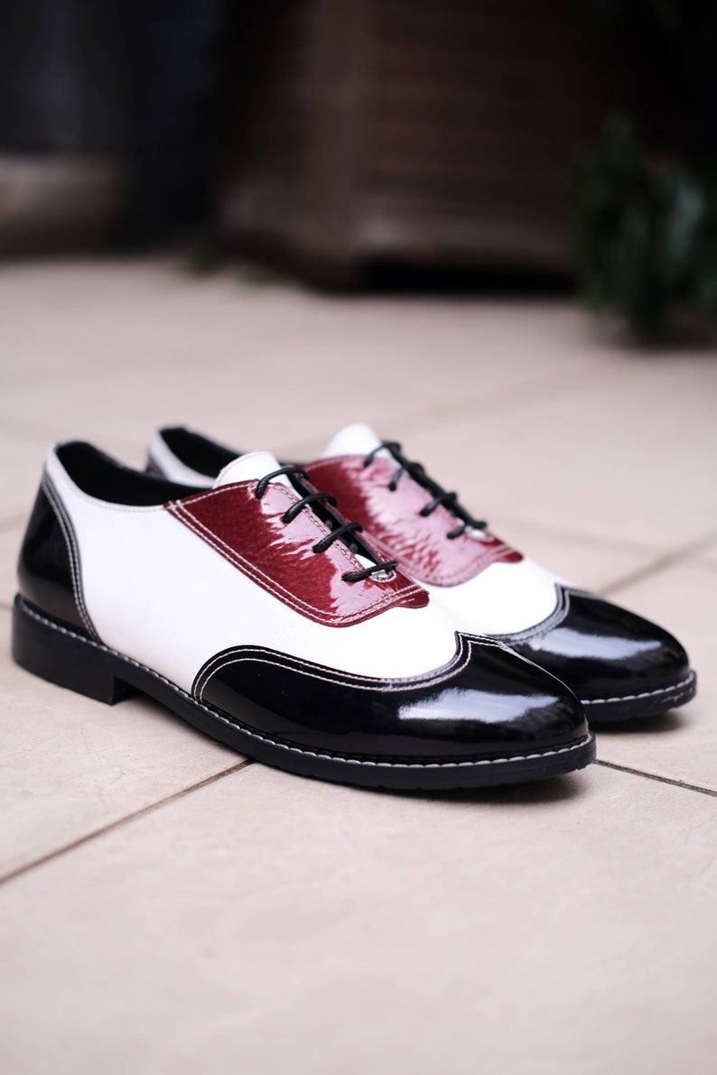 May include: A pair of black and white leather shoes with a burgundy accent. The shoes have a lace-up closure and a wingtip design.