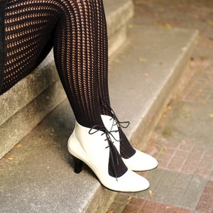 May include: A pair of white heeled boots with black lace-up detailing. The boots are worn with black fishnet tights.