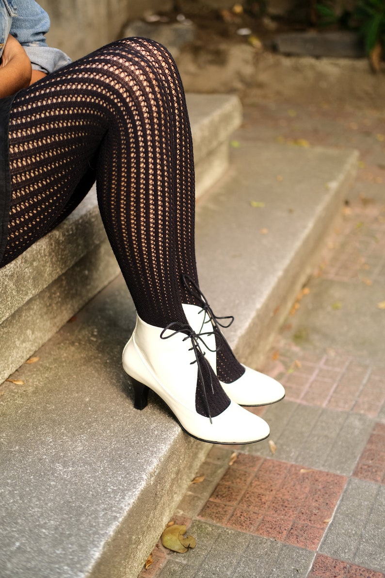 May include: A pair of white heeled boots with black lace-up detailing. The boots are worn with black fishnet tights.