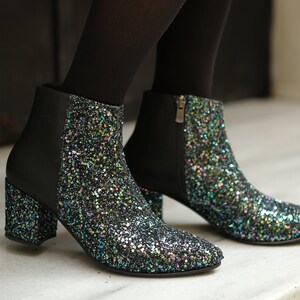 May include: Black ankle boots with a glittery, multi-colored design on the toe and heel. The boots have a block heel and a side zipper. The upper portion of the boot is black.