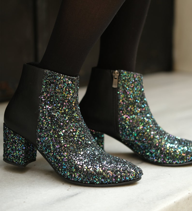 May include: Black ankle boots with a glittery, multi-colored design on the toe and heel. The boots have a block heel and a side zipper. The upper portion of the boot is black.