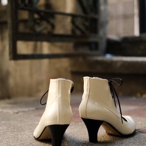 May include: A pair of white heeled ankle boots with black laces and black heels. The boots are sitting on a concrete step.