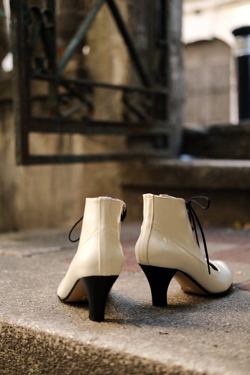 May include: A pair of white heeled ankle boots with black laces and black heels. The boots are sitting on a concrete step.