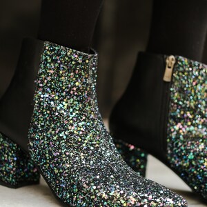 May include: Close-up of two black ankle boots with a glittery, iridescent finish. The boots have a pointed toe, a block heel, and a side zipper. The glitter is multi-colored, reflecting light.