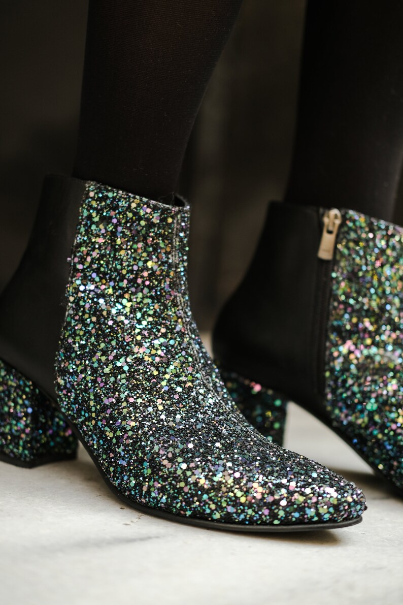 May include: Close-up of two black ankle boots with a glittery, iridescent finish. The boots have a pointed toe, a block heel, and a side zipper. The glitter is multi-colored, reflecting light.