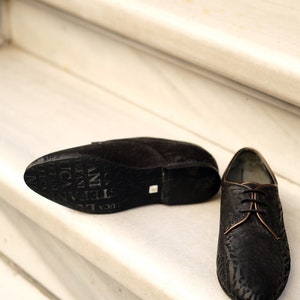 May include: A pair of black leather shoes with a lace-up fastening. The shoes have a textured pattern and a brown leather trim around the laces. The shoes are on a white marble staircase.