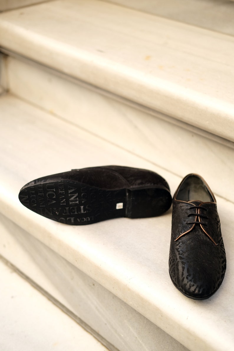 May include: A pair of black leather shoes with a lace-up fastening. The shoes have a textured pattern and a brown leather trim around the laces. The shoes are on a white marble staircase.