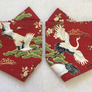 May include: Two red fabric face masks with black elastic ear loops. Each mask features a white crane design with black accents on a red background with a gold geometric pattern.