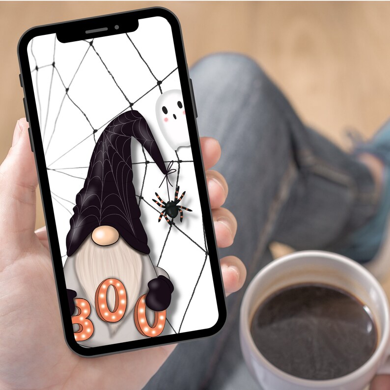 Peut inclure: Un smartphone affiche une illustration sur le th&egrave;me d'Halloween. Elle pr&eacute;sente un gnome portant un chapeau de sorci&egrave;re noir, un fant&ocirc;me et une araign&eacute;e sur une toile d'araign&eacute;e. Le mot "BOO" est &eacute;crit en lettres orange. Le t&eacute;l&eacute;phone est tenu dans une main.