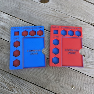 May include: Two blue and red plastic game trays with hexagonal spaces for dice and a space for a card deck. The trays are labeled "Commander DMG" and "Command Zone".