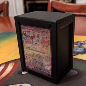 Peut inclure: Un porte-cartes noir avec une fenêtre en plastique transparente affichant une carte Pokémon représentant Sylveon, un Pokémon de type fée rose avec une capacité de charme magique. Le texte de la carte indique "Charme Magique" et "Angélite".
