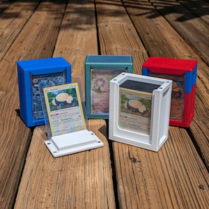 Op de afbeelding: Drie 3D-geprinte kaartspellen, een blauwe, een groene en een rode, met een witte kaarthouder in het midden. De kaarthouder heeft een transparant plastic venster om een Pokémon-kaart te tonen. De kaarten in de dozen zijn Snorlax, Gardevoir en Charizard.