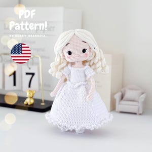 Op de afbeelding: Een handgemaakte witte gehaakte pop met krullend blond haar en een witte jurk. De pop heeft zwarte ogen en roze wangen. De afbeelding bevat de tekst "PDF Pattern!" en een Amerikaanse vlag.