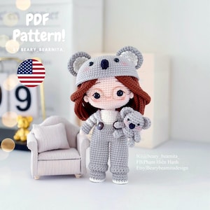 May include: A crocheted amigurumi doll wearing a koala hat and holding a smaller koala amigurumi. The doll has brown hair, glasses, and is wearing a grey jumpsuit. The doll is standing on a white surface.