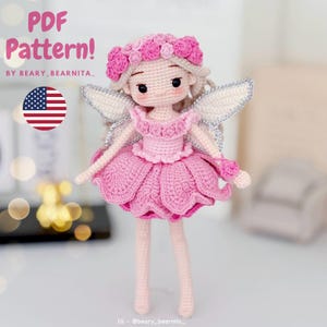 Puede incluir: Una muñeca de ganchillo con un vestido rosa de falda en forma de pétalos, una corona de flores a juego y alas blancas con borde plateado. El texto PDF Pattern! BY BEARY_BEARNITA_ y una bandera de EE. UU. aparecen en la esquina superior.