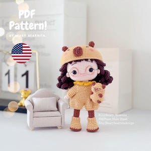 Puede incluir: Muñeca de ganchillo hecha a mano con un atuendo de oso, que incluye un sombrero y un mono, sosteniendo un pequeño oso. La muñeca tiene cabello rizado oscuro, gafas y un cuello amarillo. La imagen incluye el texto "PDF Pattern!"