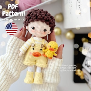 Könnte beinhalten: Häkelanleitung für eine Amigurumi-Puppe eines Jungen mit braunen Haaren und einer gelben Ente. Die Puppe trägt ein gelb-weiß gestreiftes Hemd, eine gelbe Latzhose und gelbe Schuhe. Die Ente hält eine gelbe Blume.