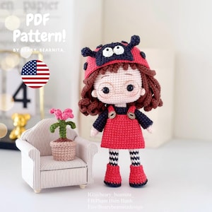 Pode incluir: Uma boneca de crochê em forma de joaninha usando um vestido vermelho, leggings listradas preto e branco e botas vermelhas. A boneca tem cabelo castanho, olhos pretos e óculos. A boneca está sentada em um sofá em miniatura com uma pequena planta em vaso.