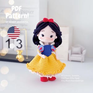 Op de afbeelding: Gehaakte Sneeuwwitje pop met een gele jurk, een blauwe bovenkant en rode schoenen. De pop heeft zwart haar en een rode strik. De pop staat op een wit oppervlak.