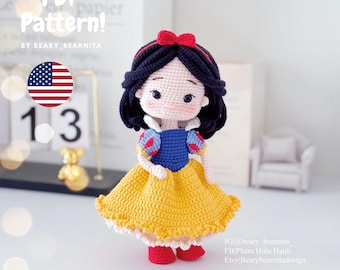 May include: Crocheted Snow White doll with a yellow dress, blue bodice, and red shoes. The doll has black hair and a red bow. The doll is standing on a white surface.