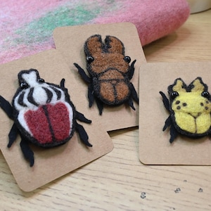 May include: Three handmade felt insect brooches on brown cards. One has a white and red design, another is brown, and the third is yellow. Each has black legs and antennae. The background includes pink and green felt.