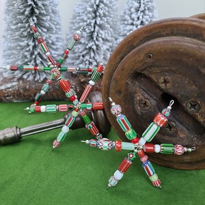 Puede incluir: Tres adornos navideños hechos a mano con forma de copo de nieve. Cada copo de nieve está hecho con cuentas de vidrio rojas, verdes, blancas y azules. Los copos de nieve están colgados de una superficie de madera.