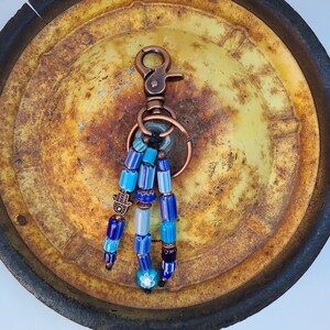 Puede incluir: Un llavero de cuentas azules y blancas con un dije de hamsa de cobre y un anillo de llave de cobre. El llavero está colgado en una superficie de metal oxidado.