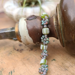 Puede incluir: Un brazalete de cuentas con cuentas verdes, blancas y moradas. Las cuentas están dispuestas en un patrón de flores y rayas.