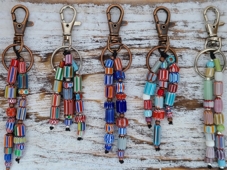 Puede incluir: Cinco llaveros de cuentas coloridas con cierres de cobre y plata. Cada llavero presenta una variedad de cuentas de vidrio de diferentes colores y patrones, incluyendo rojo, azul, verde, amarillo y blanco.