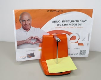 May include: An orange plastic memo holder with a metal rod and a yellow sticky note. The holder is in front of a poster with the text "ישראכרט" and a man making a heart shape with his hands.