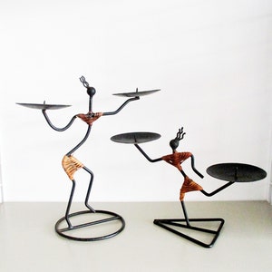 May include: Two black metal candle holders shaped like stylized figures. The figures are holding round candle plates. The figures are decorated with brown woven material.