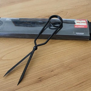 Könnte beinhalten: Schwarze Metallscheren mit einem einzigartigen Design, verpackt in einer schwarzen Box mit dem Text "EMBRO Scissors" und "HOMESS" darauf gedruckt. Die Box hat auch einen Aufkleber mit der Aufschrift "SELFRIDGES".