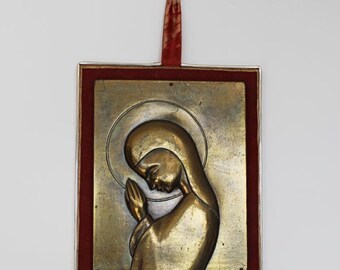 May include: A gold-toned metal plaque with a raised image of a woman in prayer. The plaque is framed in a silver-coloured frame with a red velvet backing. The plaque is hanging from a red ribbon.