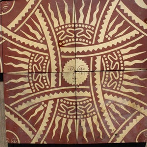 Może przedstawiać: Zestaw płytek w kolorze terakoty z powtarzającym się wzorem słońca. Słońce ma twarz pośrodku, otoczoną promienistymi liniami i dekoracyjnymi wzorami w kolorze kremowym. Płytki są ułożone razem, prezentując pełny wzór.