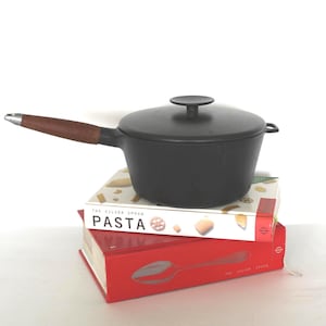 以下が含まれることがあります： 蓋と木製の持ち手が付いた黒い鋳鉄製の片手鍋が、2冊の料理本の上に置かれています。料理本のタイトルは「Pasta」と「The Silver Spoon」です。片手鍋は濃いグレー色です。