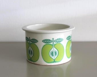 May include: A small, off-white ceramic container with a rounded rim. The container is decorated with a repeating pattern of green apple halves with white and green details. The design is a vintage style.